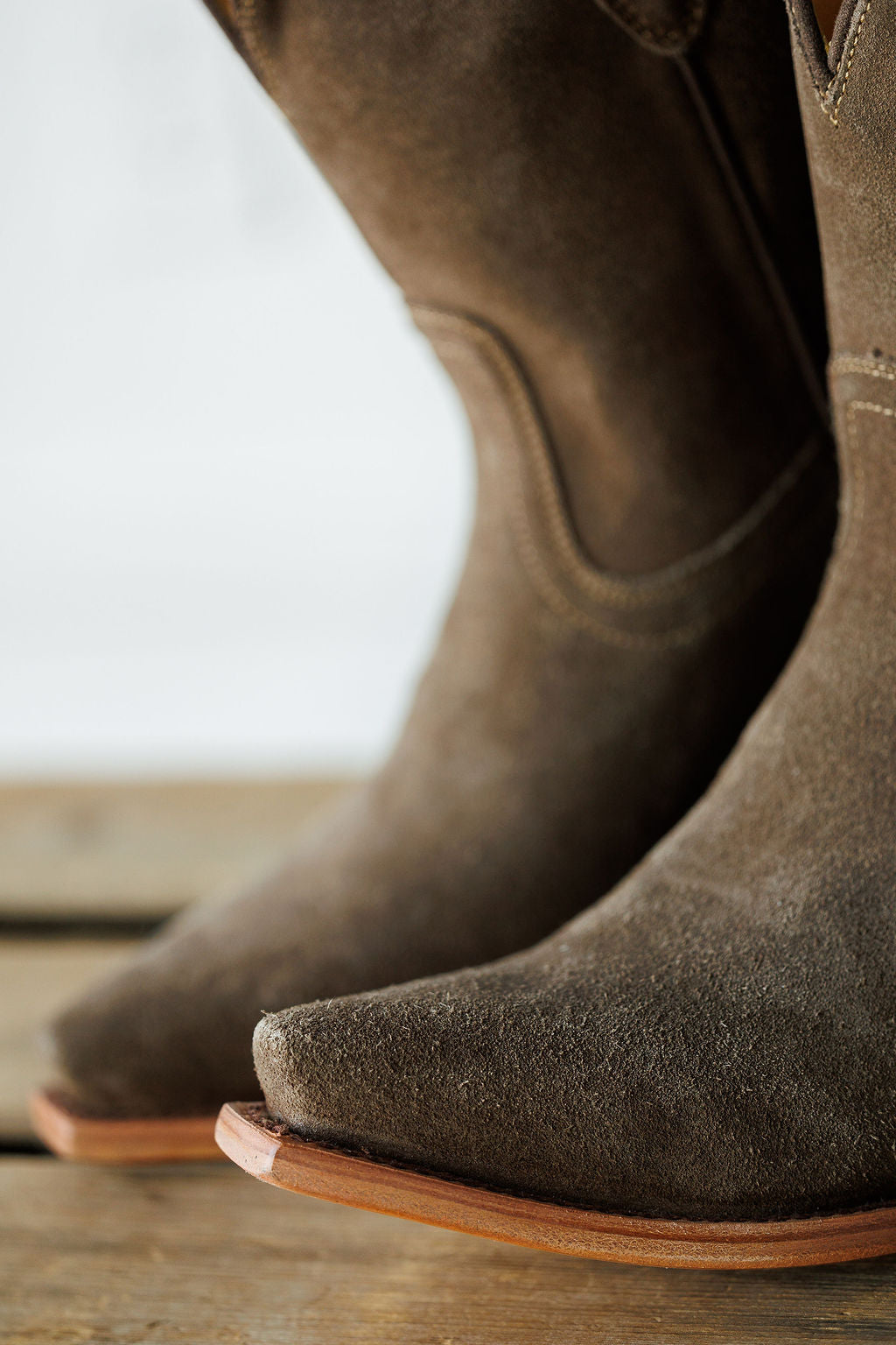 Close-up of the toes of The Cordova cowboy boots by Savannah Sevens western life{&}style, featuring brown rough out leather, light brown leather soles, fine stitching, and a slightly distressed texture on a wooden surface. Made by Black Star. 