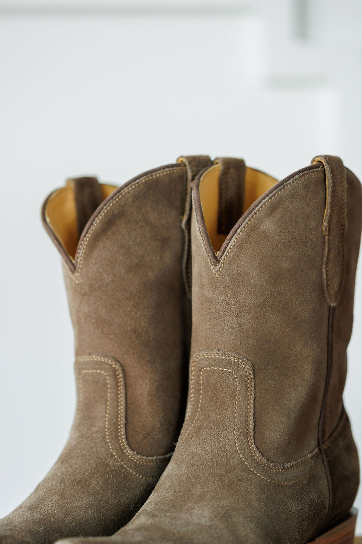 A close-up of The Cordova booties by Savannah Sevens western life{&}style, featuring brown rough out leather, decorative stitching, and pull straps, set against a softly blurred background. Made by Black Star. 