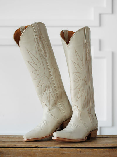 A pair of The Loretta Boot {Cream} by Savannah Sevens western life{&}style, tall cowboy boots in light beige with decorative stitching and premium cowhide leather, stand upright on a wooden surface against a white paneled background. Boots handmade by Tecovas.