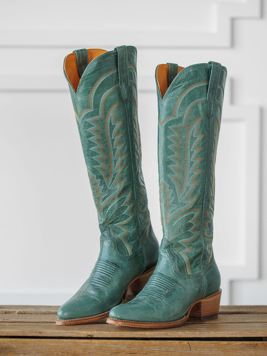 A pair of tall, sagebrush-green "The Abby Boot" by Savannah Sevens western life{&}style with decorative stitching and wooden heels, displayed upright on a wooden surface against a white paneled background. Boots handmade by Tecovas.