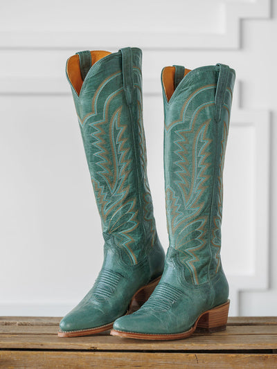 A pair of tall, sagebrush-green "The Abby Boot" by Savannah Sevens western life{&}style with decorative stitching and wooden heels, displayed upright on a wooden surface against a white paneled background. Boots handmade by Tecovas.