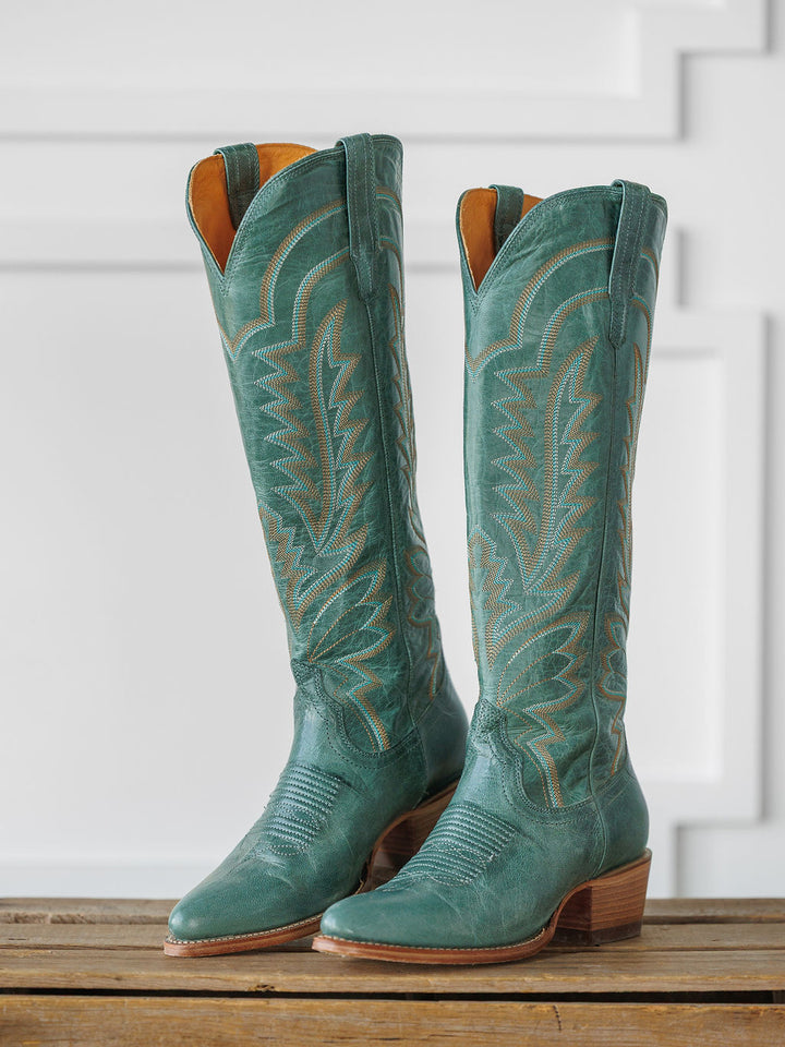 A pair of tall, sagebrush-green "The Abby Boot" by Savannah Sevens western life{&}style with decorative stitching and wooden heels, displayed upright on a wooden surface against a white paneled background. Boots handmade by Tecovas.