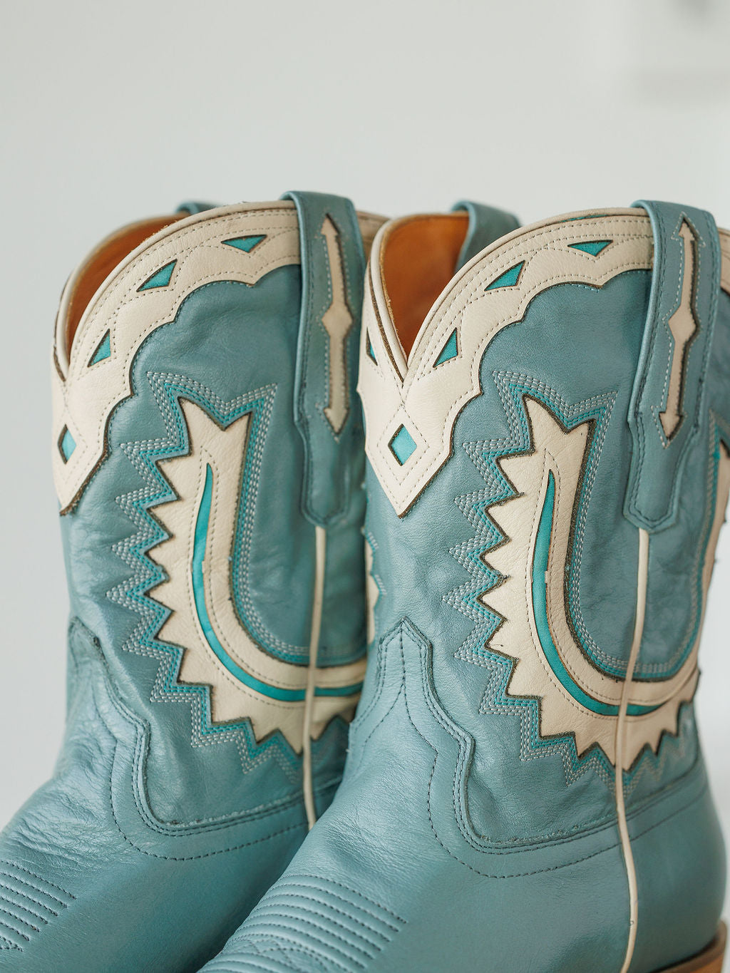 A close-up of The Jolene Boot {Seafoam} by Savannah Sevens western life{&}style, showing turquoise cowhide with cream and teal stitching and patterns that emphasize the retro western design and detailed craftsmanship on the uppers. Boots handmade by Tecovas.