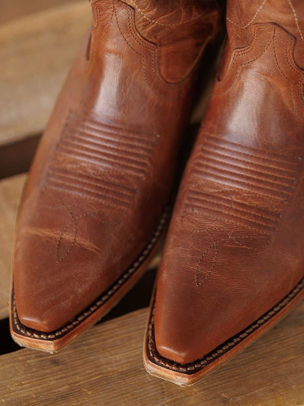Close-up of the pointed toes of The Annie Boot {Scotch} by Savannah Sevens western life{&}style, featuring brown leather with decorative stitching and textured detailing, resting on a wooden surface. Boots handmade by Tecovas.