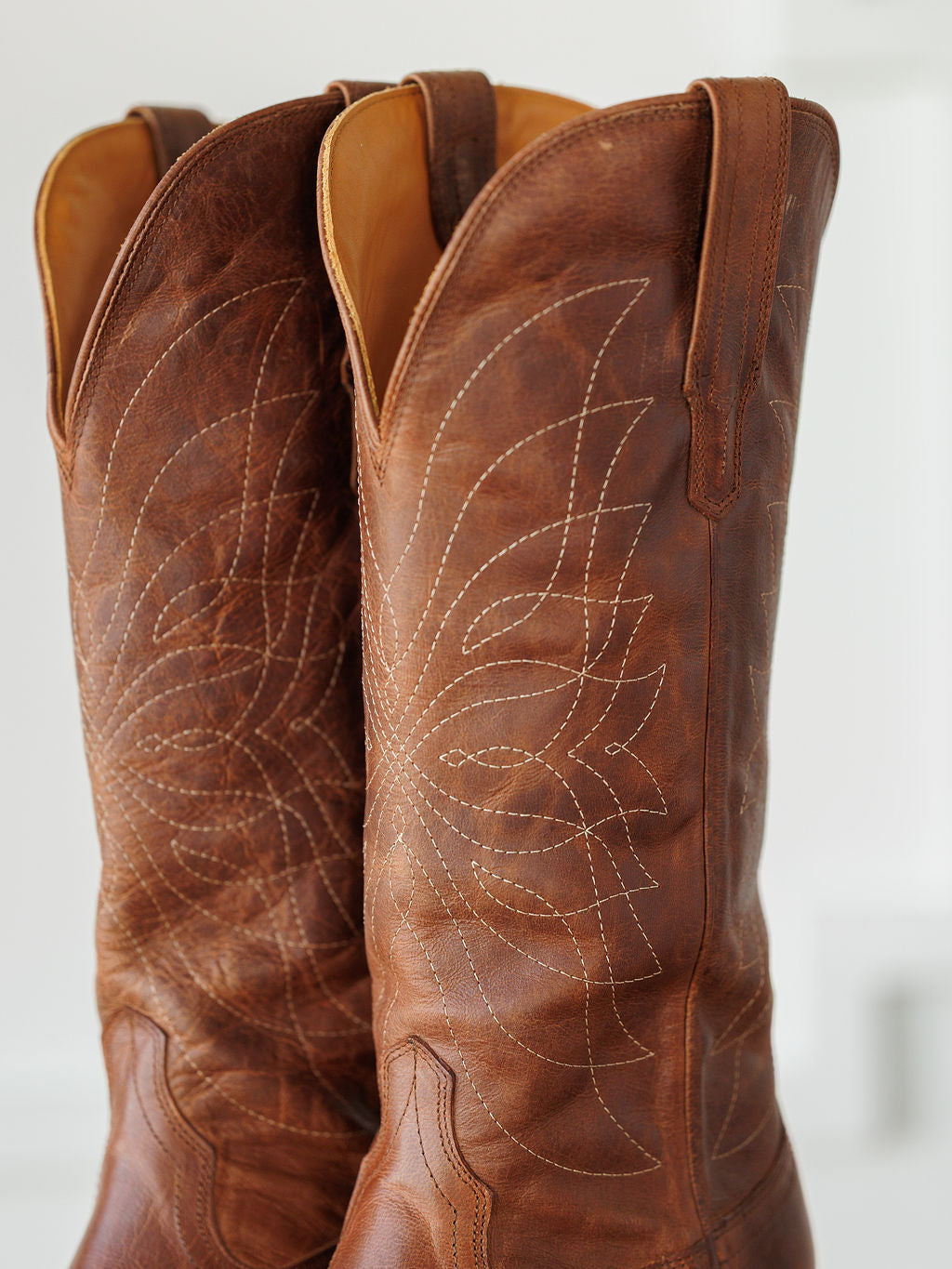 A close-up of The Annie Boot {Scotch} by Savannah Sevens western life{&}style highlights brown leather boots with swirling white stitching, pull straps, and a snip toe design against a softly blurred backdrop. Boots handmade by Tecovas.