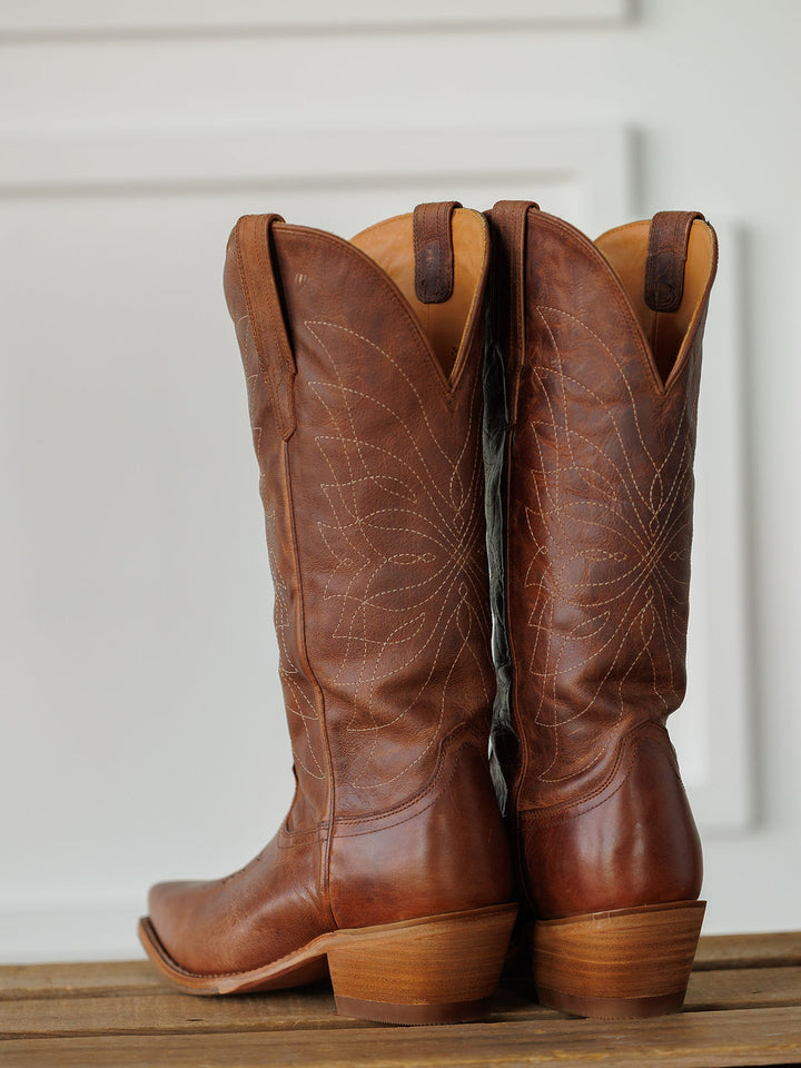 A pair of The Annie Boot {Scotch} by Savannah Sevens western life{&}style, featuring brown leather and decorative stitching, stand upright on a wooden surface against a white background. Boots handmade by Tecovas.