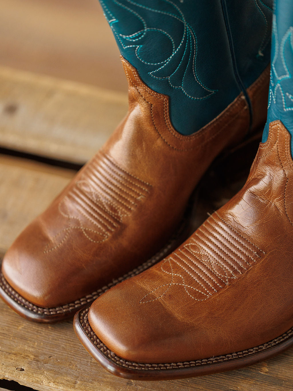 A close-up of The Maggie Boot {Teal} by Savannah Sevens western life{&}style, featuring premium goat leather and teal stitching on the upper shaft, resting on a wooden surface. Boots handmade by Tecovas.