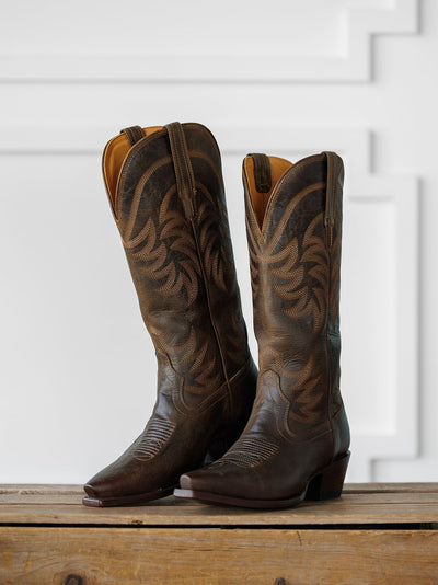 The Annie Boot {Cafe} by Savannah Sevens western life{&}style, a tall dark brown leather snip toe boot with decorative stitching, stands on a wooden surface before a white paneled background. Boots handmade by Tecovas.