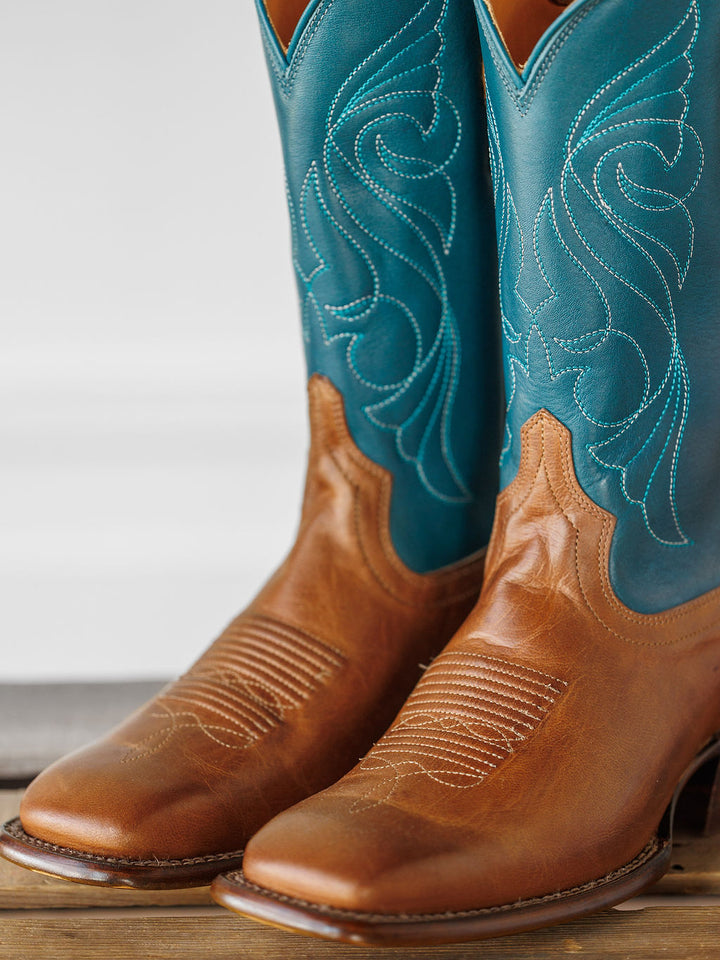 The Maggie Boot {Teal} by Savannah Sevens western life{&}style features premium goat leather: a light brown foot, vibrant teal shafts with white stitching, displayed on a wooden surface. Boots handmade by Tecovas.