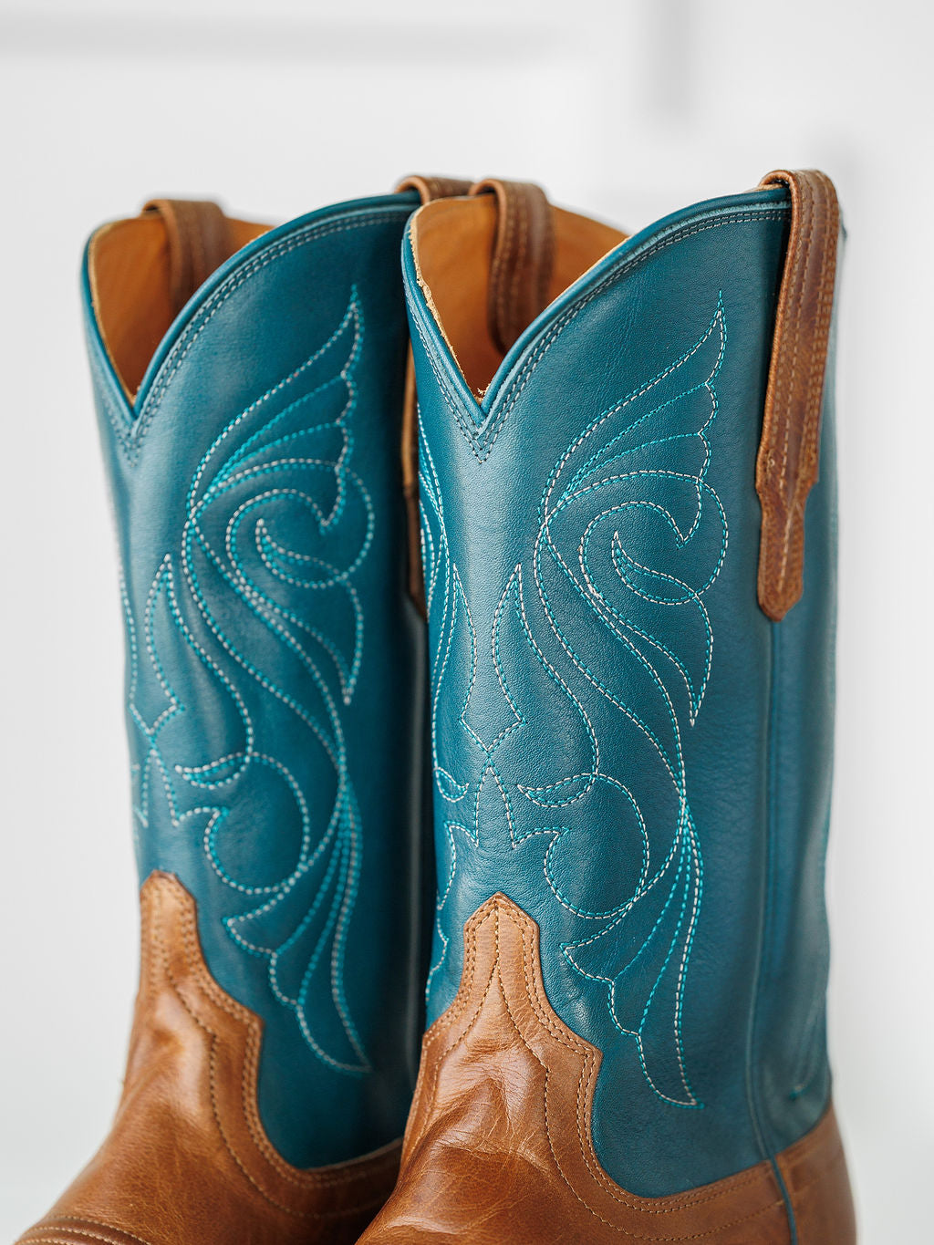 A close-up of The Maggie Boot {Teal} by Savannah Sevens western life{&}style shows blue shafts, decorative stitching, and premium goat leather in a square toe Western design, set against a softly blurred light background. Boots handmade by Tecovas.