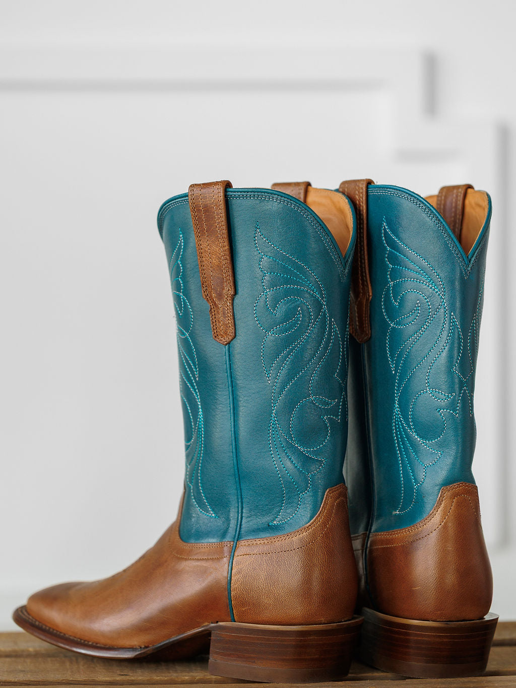 A pair of Savannah Sevens western life{&}style The Maggie Boot {Teal} with brown premium goat leather feet and teal shafts with white stitching, displayed side by side on a wooden surface against a light background. Boots handmade by Tecovas.