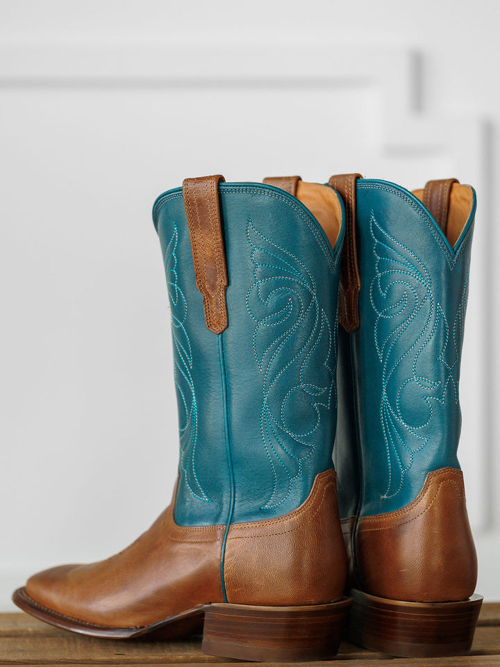A pair of Savannah Sevens western life{&}style The Maggie Boot {Teal} with brown premium goat leather feet and teal shafts with white stitching, displayed side by side on a wooden surface against a light background. Boots handmade by Tecovas.