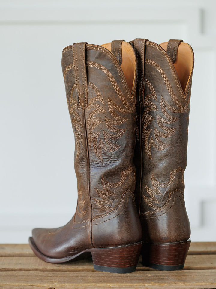 The Annie Boot {Cafe} by Savannah Sevens western life{&}style, crafted from premium goat leather with decorative stitching and a snip toe, are angled on a wooden surface against a light background. Boots handmade by Tecovas.