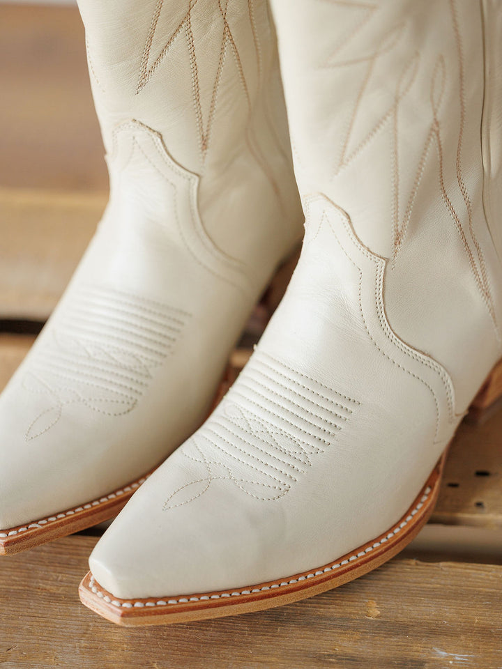 Close-up of The Loretta Boot {Cream} by Savannah Sevens western life{&}style—stylish white cowboy boots with decorative stitching, pointed toes, tan soles, and premium cowhide leather, set on a wooden surface. Boots handmade by Tecovas.