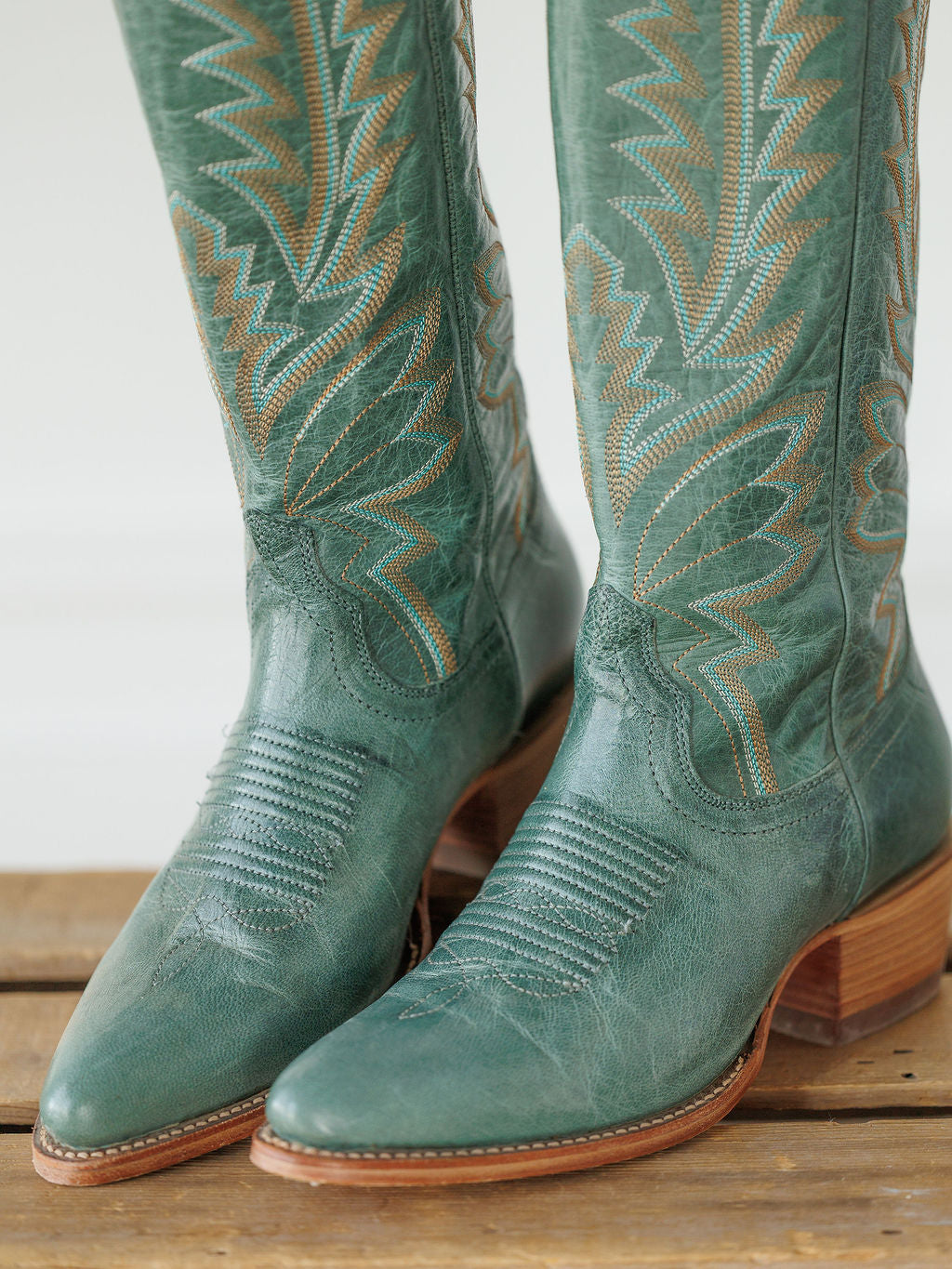 A close-up of The Abby Boot {Sagebrush} by Savannah Sevens western life{&}style—tall, green Western leather boots with intricate stitching, pointed toes, and wooden heels—standing on a wooden surface. Boots handmade by Tecovas.