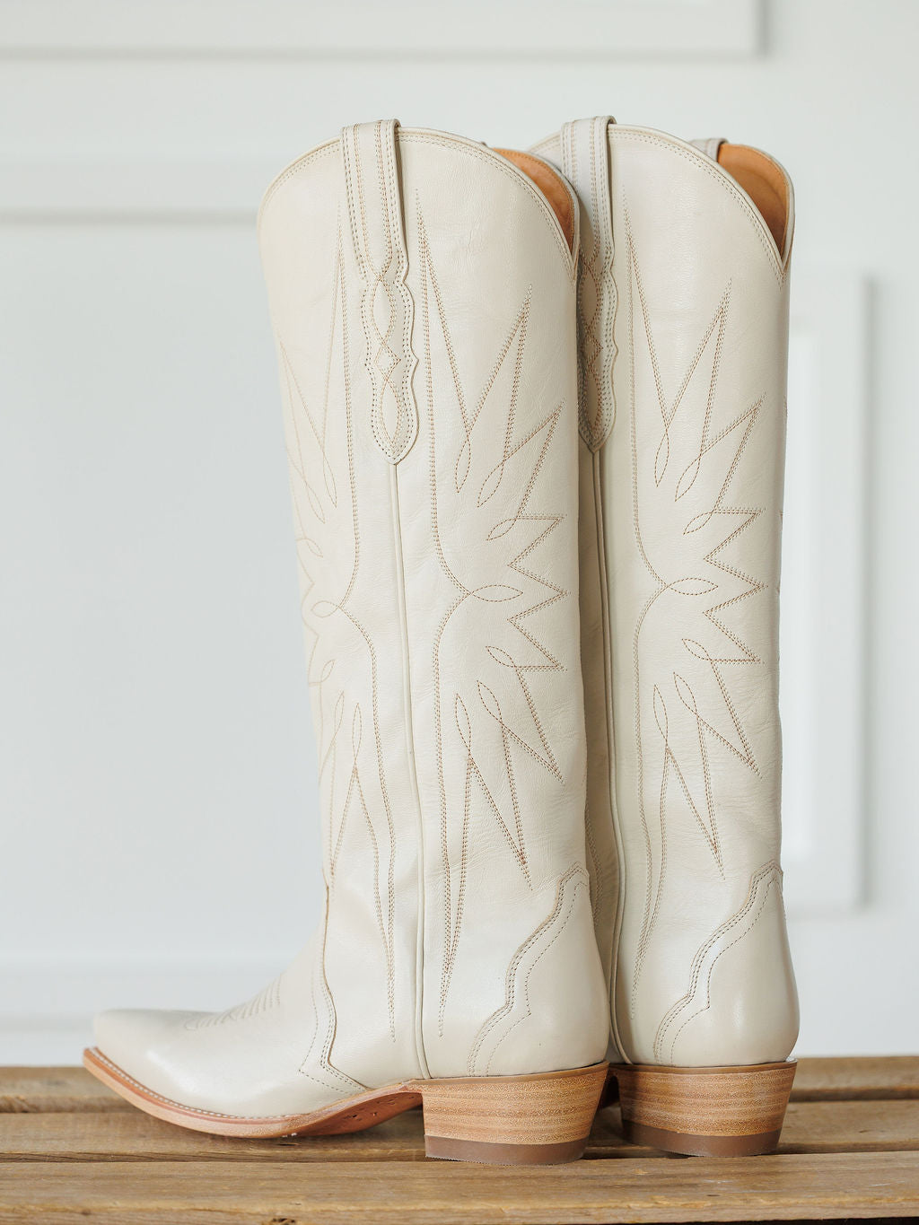 A pair of The Loretta Boot {Cream} by Savannah Sevens western life{&}style—tall, white premium cowhide leather cowboy boots with light brown heels and decorative stitching—stand side by side on a wooden surface against a white background. Boots handmade by Tecovas.