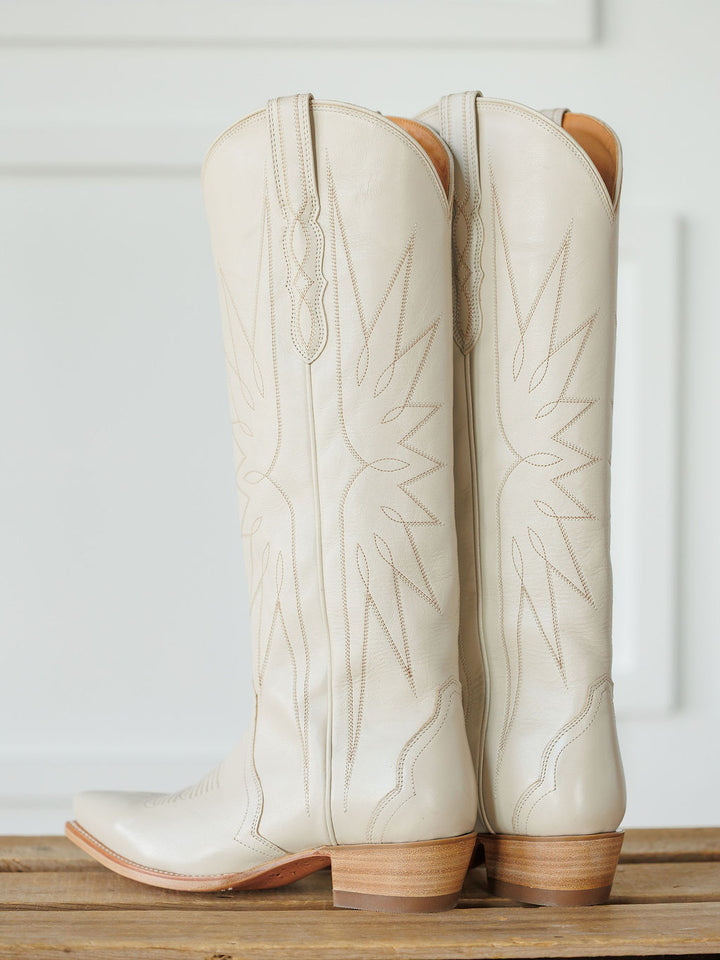 A pair of The Loretta Boot {Cream} by Savannah Sevens western life{&}style—tall, white premium cowhide leather cowboy boots with light brown heels and decorative stitching—stand side by side on a wooden surface against a white background. Boots handmade by Tecovas.