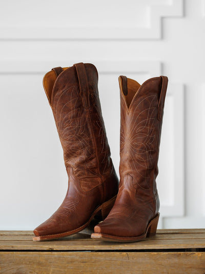 The Annie Boot {Scotch} by Savannah Sevens western life{&}style—a tall, brown Western leather boot with decorative stitching—is displayed side by side on a wooden surface against a white, textured background. Boots handmade by Tecovas.
