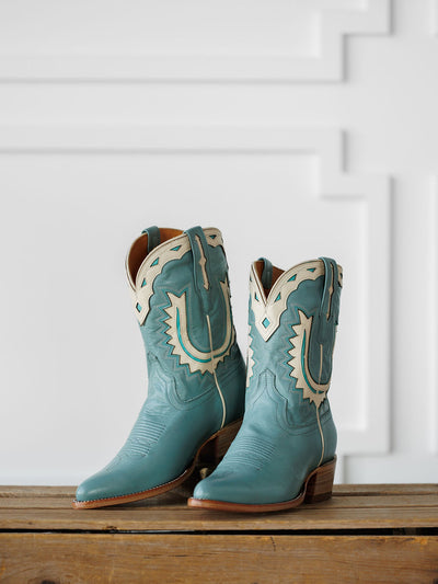 The Jolene Boot {Seafoam} by Savannah Sevens western life{&}style: retro teal leather cowboy boots with cream stitching and cutouts, shown on a wooden surface against a white geometric-patterned wall. Boots handmade by Tecovas.