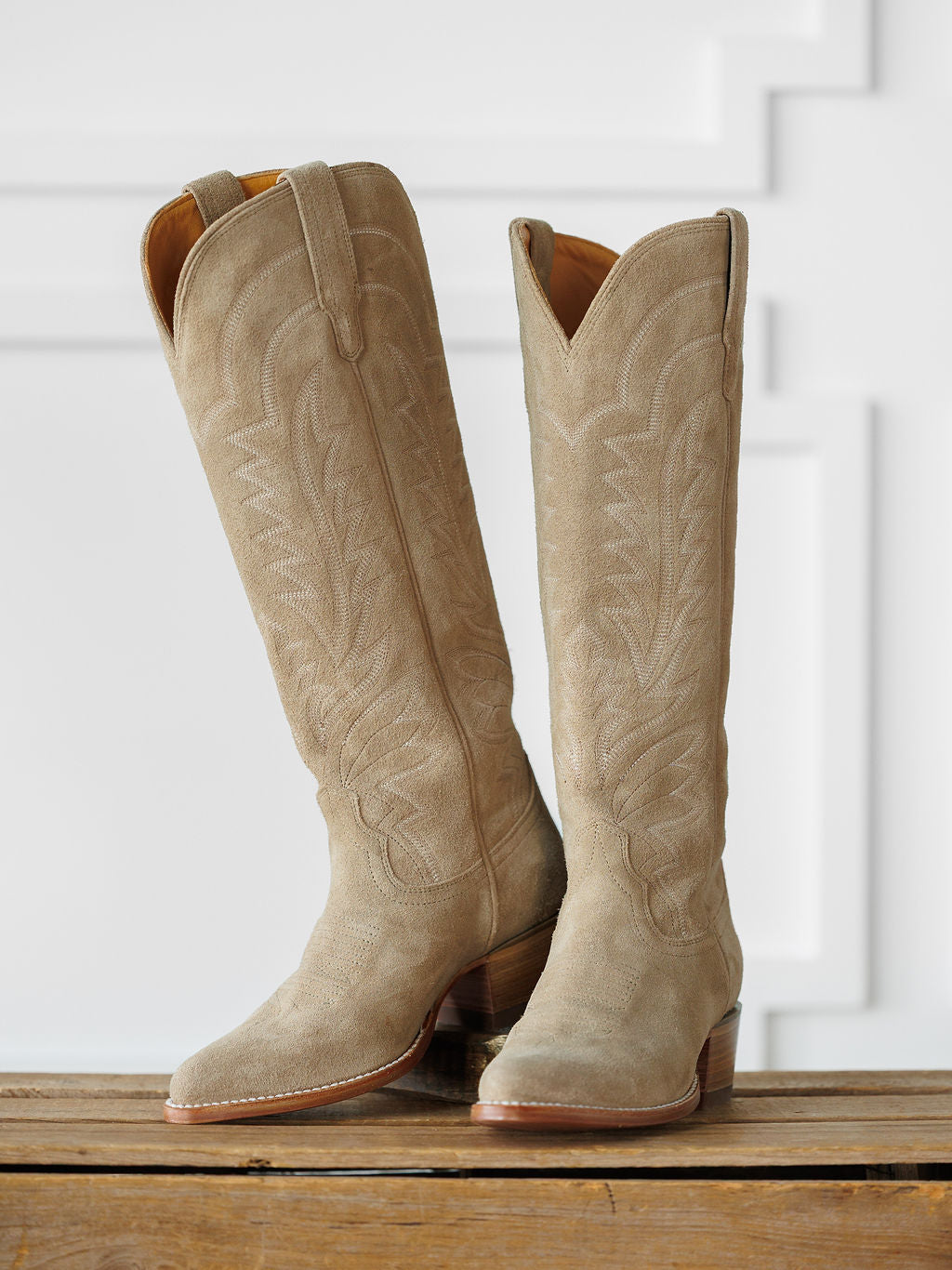 A pair of The Abby Boot {Driftwood} by Savannah Sevens western life{&}style—tall, light tan suede cowboy boots with decorative stitching—stand on a wooden surface against a white paneled background. Boots handmade by Tecovas.