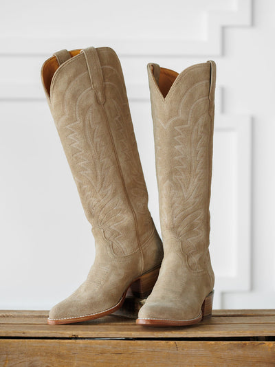 A pair of The Abby Boot {Driftwood} by Savannah Sevens western life{&}style—tall, light tan suede cowboy boots with decorative stitching—stand on a wooden surface against a white paneled background. Boots handmade by Tecovas.