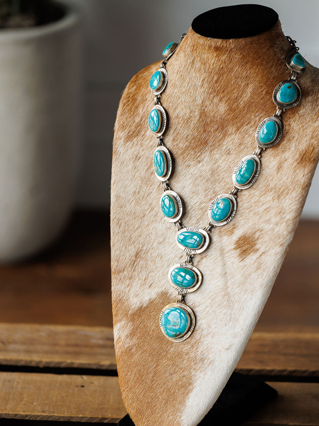 The Nogales Necklace by Savannah Sevens western life{&}style, featuring large Kingman turquoise stones, is showcased on a brown and white cowhide stand atop a wooden surface with a blurred plant pot in the background.