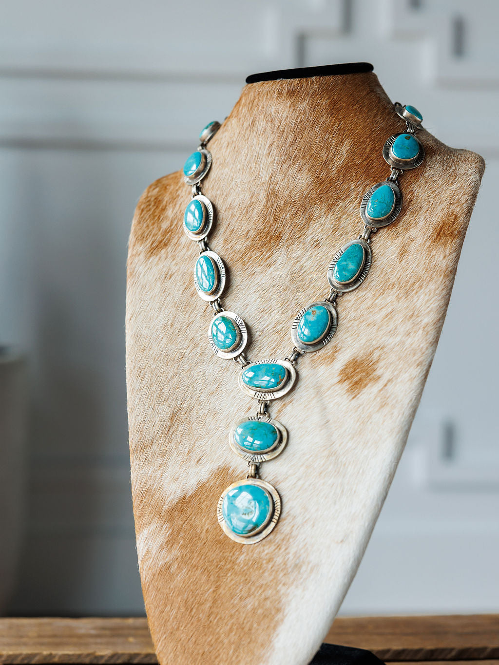 The Nogales Necklace by Savannah Sevens western life{&}style features handmade Kingman turquoise and sterling silver oval stones, displayed on a brown and white cowhide stand with a softly blurred background.
