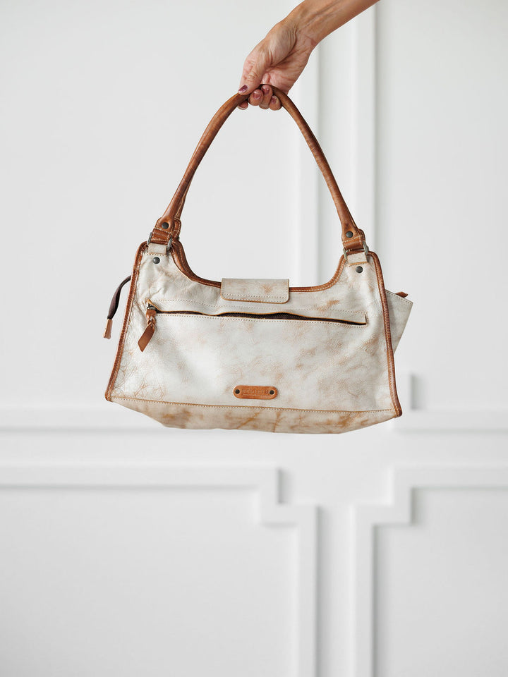 A hand holds up The Rowley by Savannah Sevens western life{&}style, a beige and brown full grain leather handbag with a front zipper pocket, top flap, and double handles, against a white wall with decorative molding.