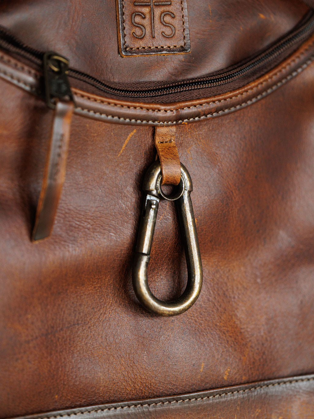 Close-up of the Waynesboro Backpack by Savannah Sevens western life{&}style, featuring worn full grain leather, an embossed logo patch above a zippered pocket, and a metal carabiner attached below.