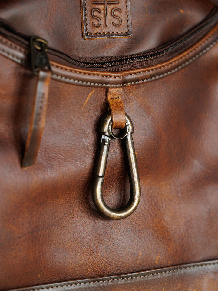 Close-up of the Waynesboro Backpack by Savannah Sevens western life{&}style, featuring worn full grain leather, an embossed logo patch above a zippered pocket, and a metal carabiner attached below.
