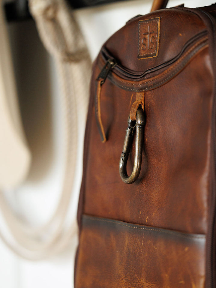 Close-up of the Waynesboro Backpack by Savannah Sevens western life{&}style, made of full grain leather. Features a metal carabiner on the zipper, visible stitching, and an embossed logo patch. A blurred rope appears in the background.