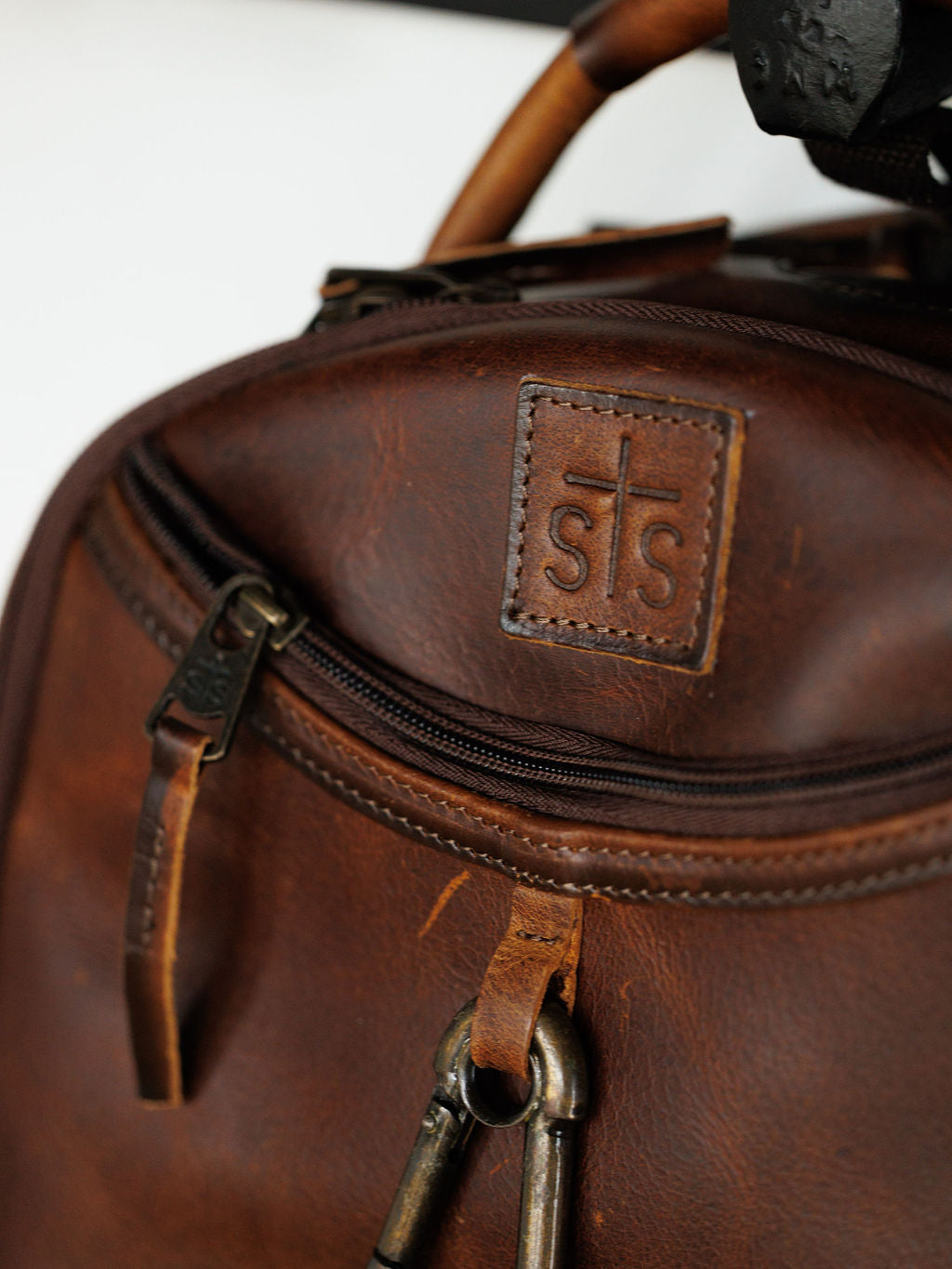 Close-up of the Waynesboro Backpack by Savannah Sevens western life{&}style, crafted from full grain leather with a front zip pocket, stitched cross logo patch with "sts," metal clips, and leather zipper pulls.