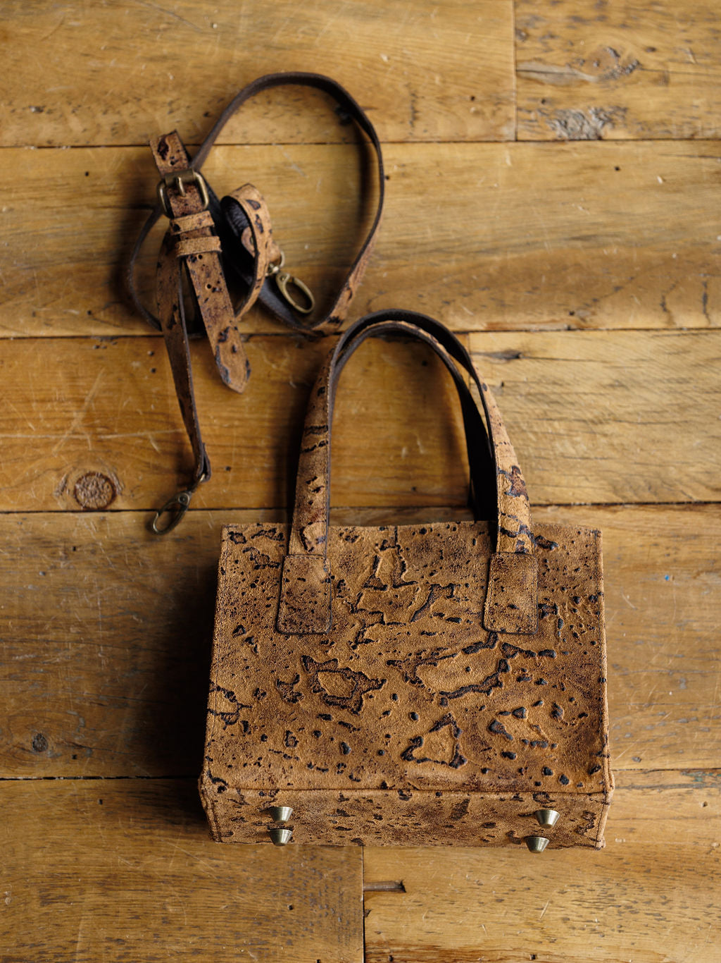 The Nubuck by Savannah Sevens western life{&}style is a brown nubuck leather handbag with short handles, a detachable shoulder strap, natural textured pattern, and metal studs on the bottom, displayed on a wooden surface.