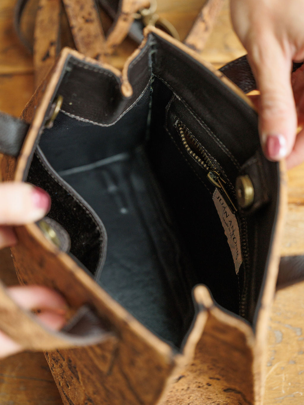 A person opens The Nubuck handbag by Savannah Sevens western life{&}style, showing its brown textured nubuck leather, black interior, zippered pocket, brass snap closures, and a Lunnamon label.