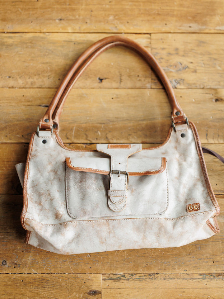 The Rowley by Savannah Sevens western life{&}style is a cream full grain leather handbag with brown trim, front buckle pocket, and double handles, shown on a wooden surface.