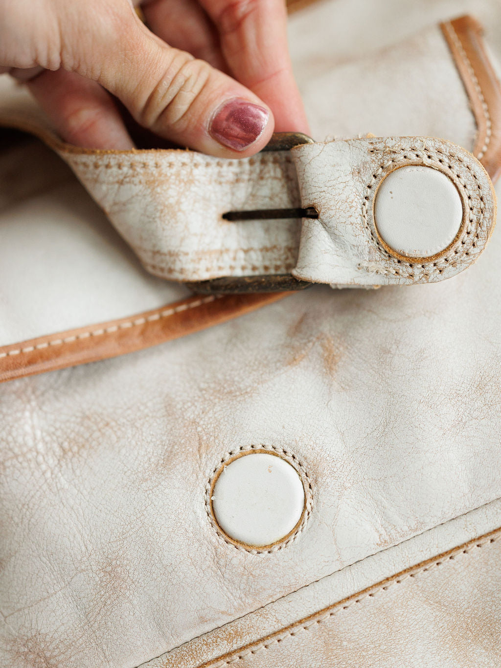 A close-up of a hand with  pulling on the strap of The Rowley by Savannah Sevens western life{&}style, a rustic full grain leather handbag with visible stitching and round button-like clasps.