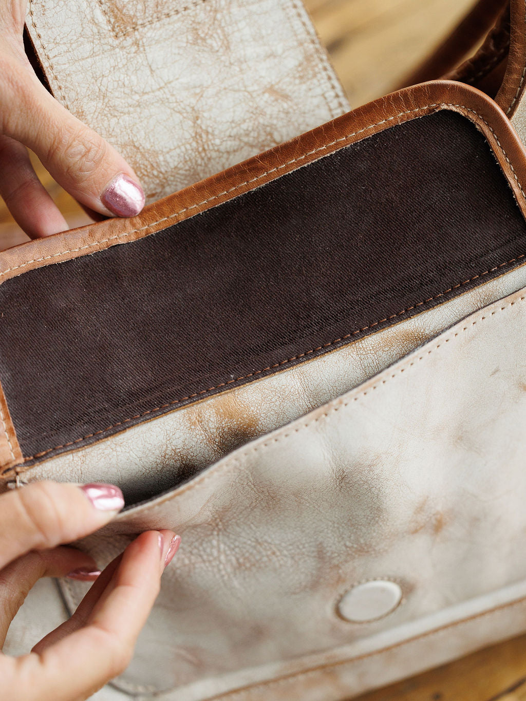 A person with metallic pink nails opens the front flap of The Rowley, a rustic leather handbag by Savannah Sevens western life{&}style, revealing its dark brown lining.