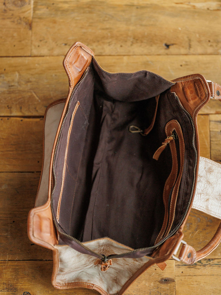 Top-down view of The Rowley by Savannah Sevens western life{&}style, a rustic full grain leather handbag with dark fabric lining and two zippered pockets, placed on a rustic wooden surface.