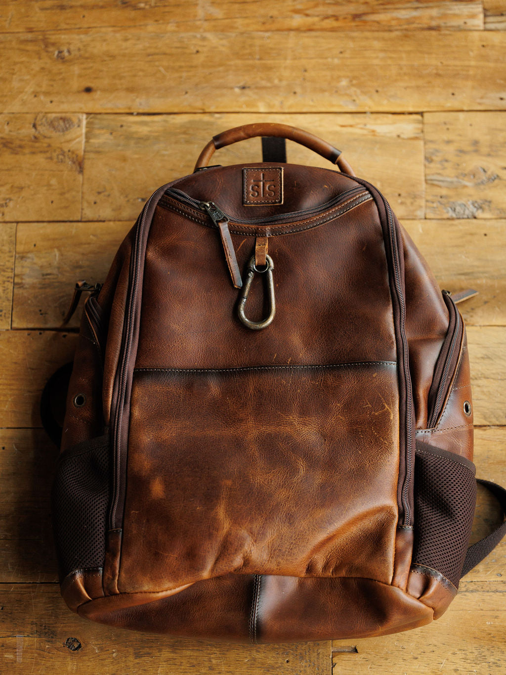 The Waynesboro Backpack by Savannah Sevens western life{&}style, crafted from brown full grain leather with a front zipper pocket, metal clasp, and side mesh pockets, features a worn vintage look and sits on a wooden floor.