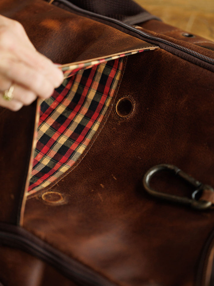 A hand lifts the zipper of the Waynesboro Backpack by Savannah Sevens western life{&}style, revealing a red, black, and tan plaid lining. A metal clasp is attached to the outside of this men's concealed carry backpack.