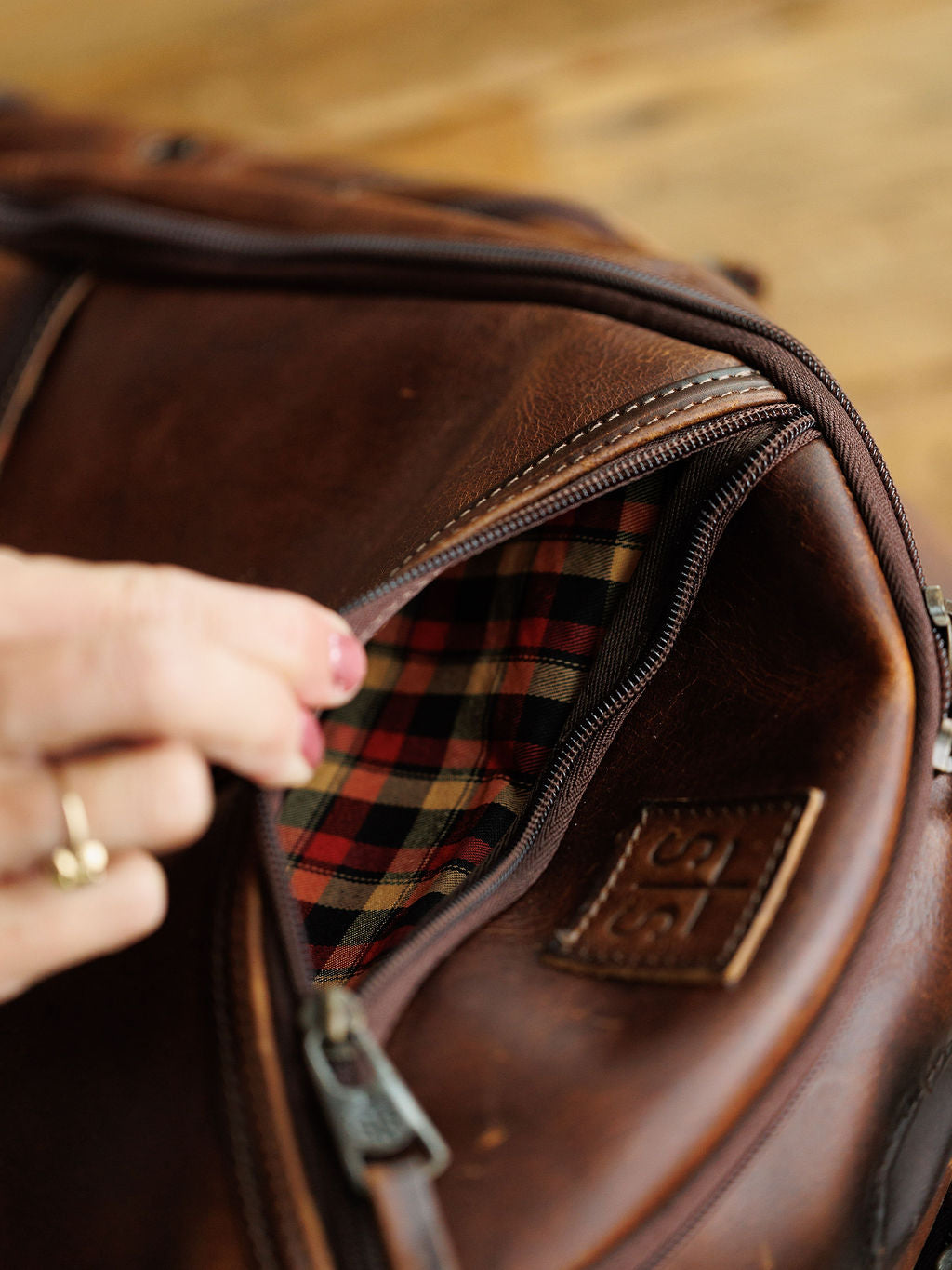 A hand with a gold ring unzips the Savannah Sevens western life{&}style Waynesboro Backpack, showing its red and black checkered lining inside the brown full grain leather exterior.