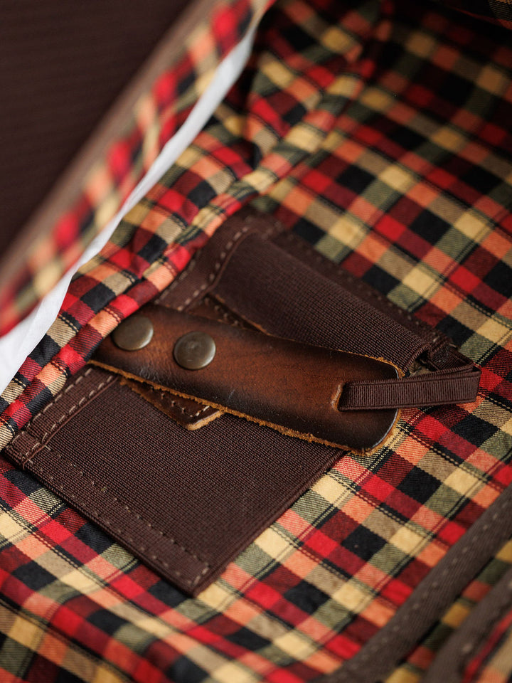 Close-up of the Waynesboro Backpack by Savannah Sevens western life{&}style, featuring full grain leather, red/yellow/black plaid lining, brown leather strap with metal rivets, and brown fabric trim inside.