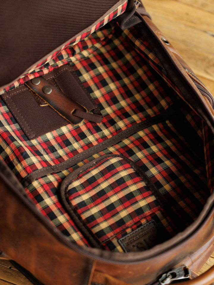The Waynesboro Backpack by Savannah Sevens western life{&}style is open to show its brown leather exterior, red, black, and beige plaid lining, multiple compartments, and an interior leather strap with a metal button.