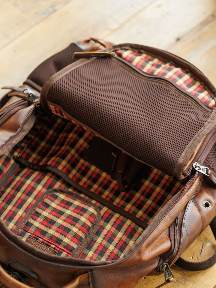 The Waynesboro Backpack by Savannah Sevens western life{&}style is shown open on a wooden floor, revealing its plaid red, yellow, and black fabric lining with visible sections and pockets.