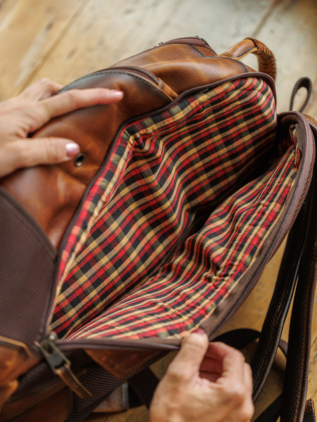 A close-up shows hands opening the Waynesboro Backpack by Savannah Sevens western life{&}style, revealing its red, yellow, and black plaid-lined interior. The full grain leather bag rests on a wooden surface.