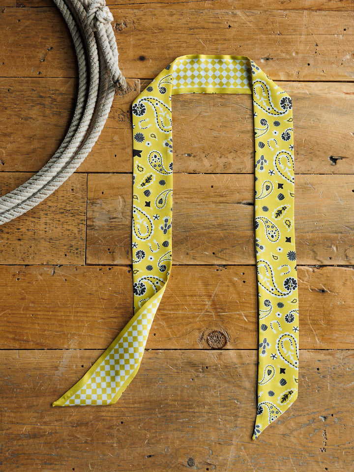 The Lucky Paisley Twilly in yellow from Savannah Sevens western life{&}style, featuring a paisley graphic and checkered design, is displayed on a rustic wooden surface beside a coiled white rope.