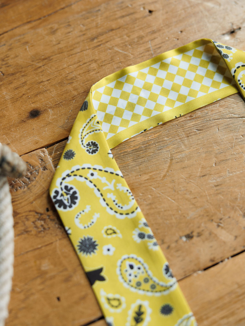 The Lucky Paisley Twilly {Yellow} by Savannah Sevens western life{&}style features a yellow chiffon scarf with black and white paisley print on one side and a yellow-and-white diamond pattern on the reverse, displayed on wood.