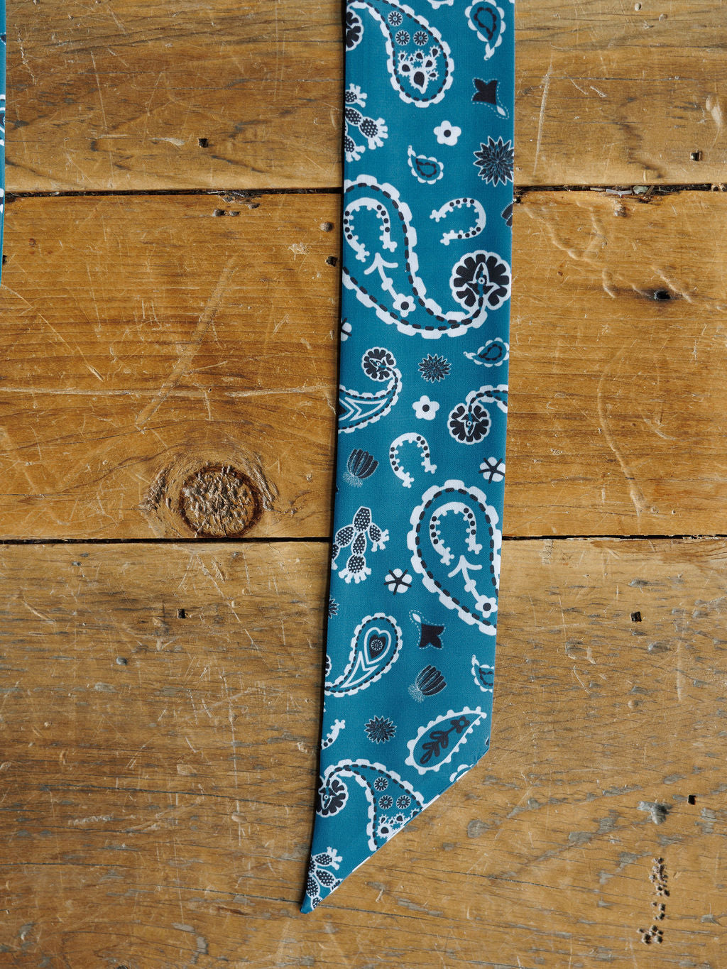 The Lucky Paisley Twilly {Cool Blue} by Savannah Sevens western life{&}style, featuring a teal paisley design with white and black accents, is displayed flat on a rustic wooden surface with visible grain and knots.