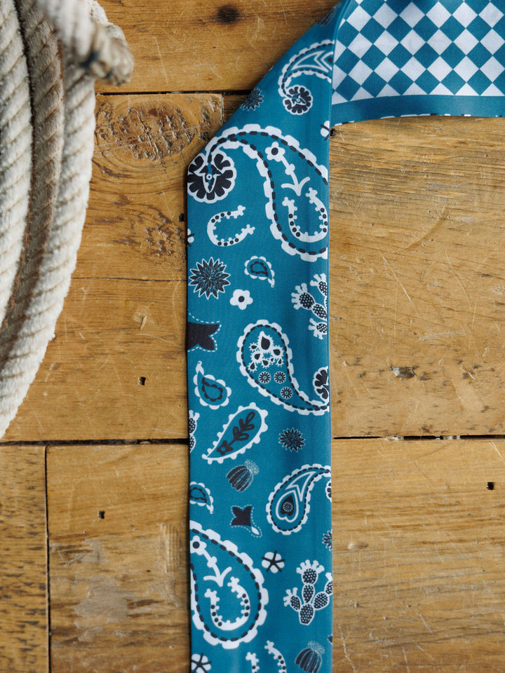 The Lucky Paisley Twilly {Cool Blue} by Savannah Sevens western life{&}style, featuring white and black paisley and floral patterns with a blue and white diamond-checkered underside, rests on wood beside a coiled rope.