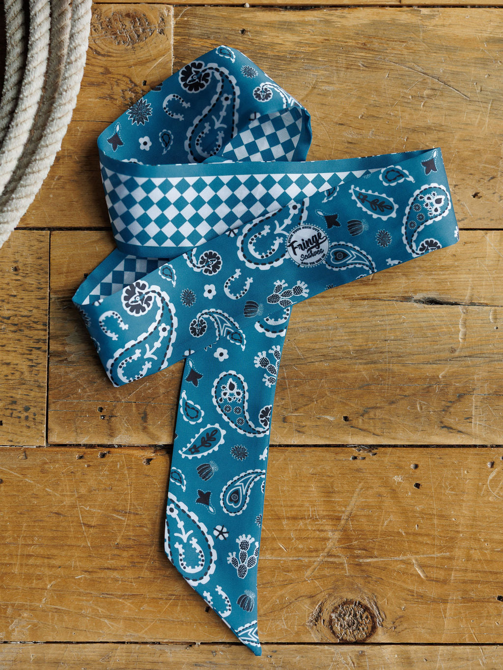 The Lucky Paisley Twilly {Cool Blue} by Savannah Sevens western life{&}style, with blue paisley print and white/black accents, is styled as a satin tie on wood. Underside features blue-white diamonds with the Friyay logo; a rope appears to the left.
